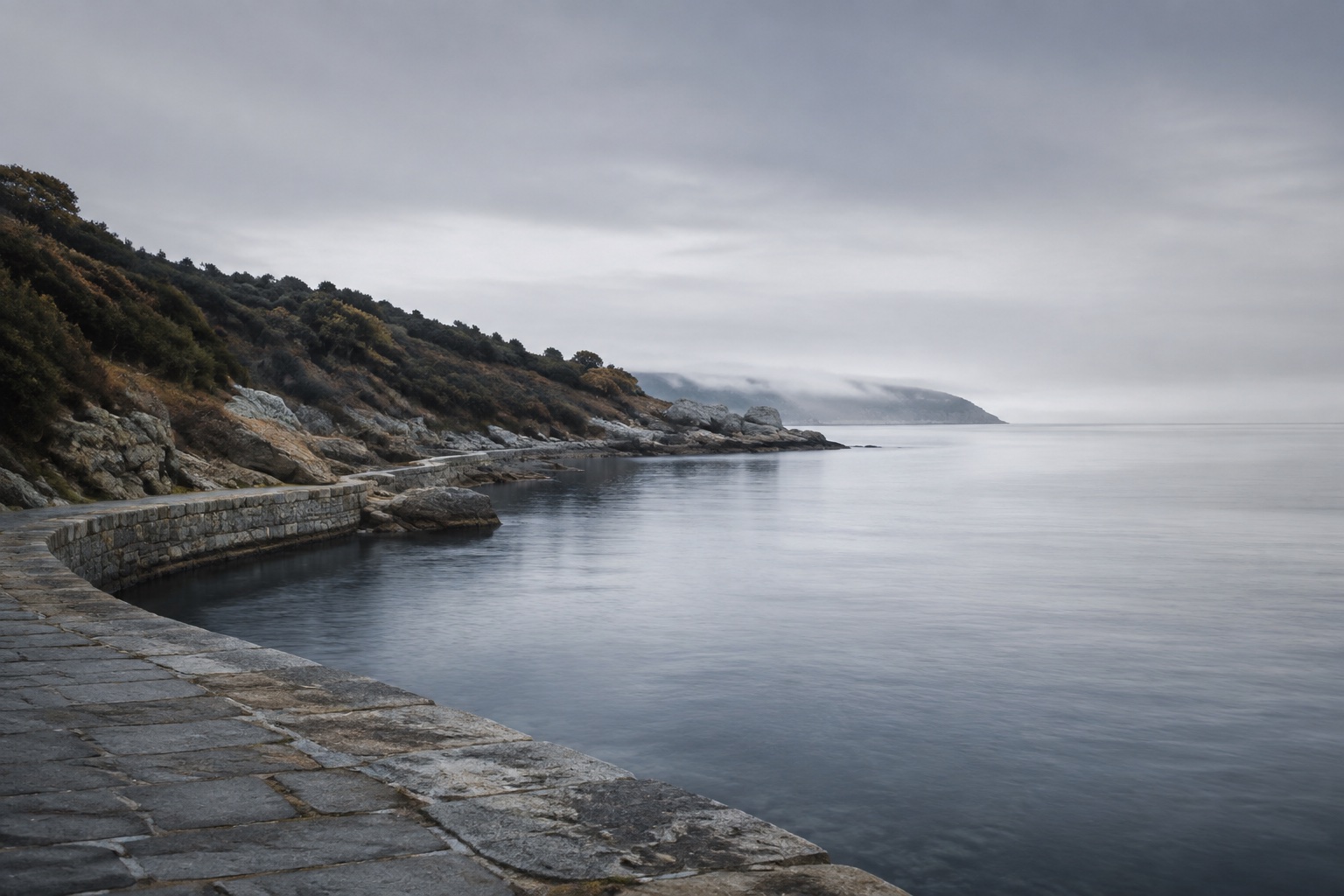 A stone promenade follows a quiet inlet around a bend softened by sea mist.